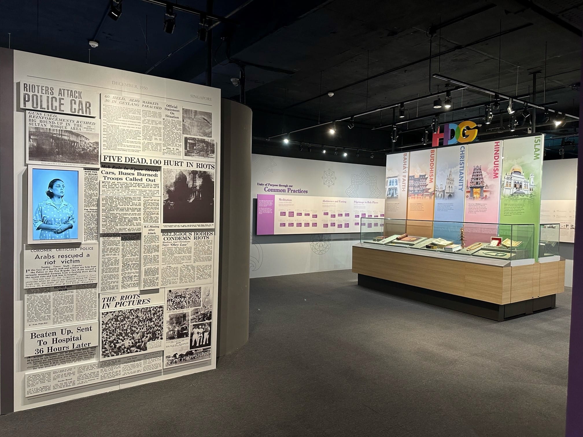 Museum exhibit with a large newspaper wall about 1950 Singapore riots and a display on world religions including Islam, Hinduism, Christianity, Buddhism.
